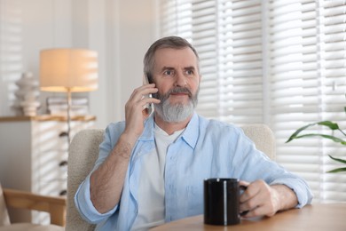 Photo of Senior man with cup of drink talking on smartphone at table indoors