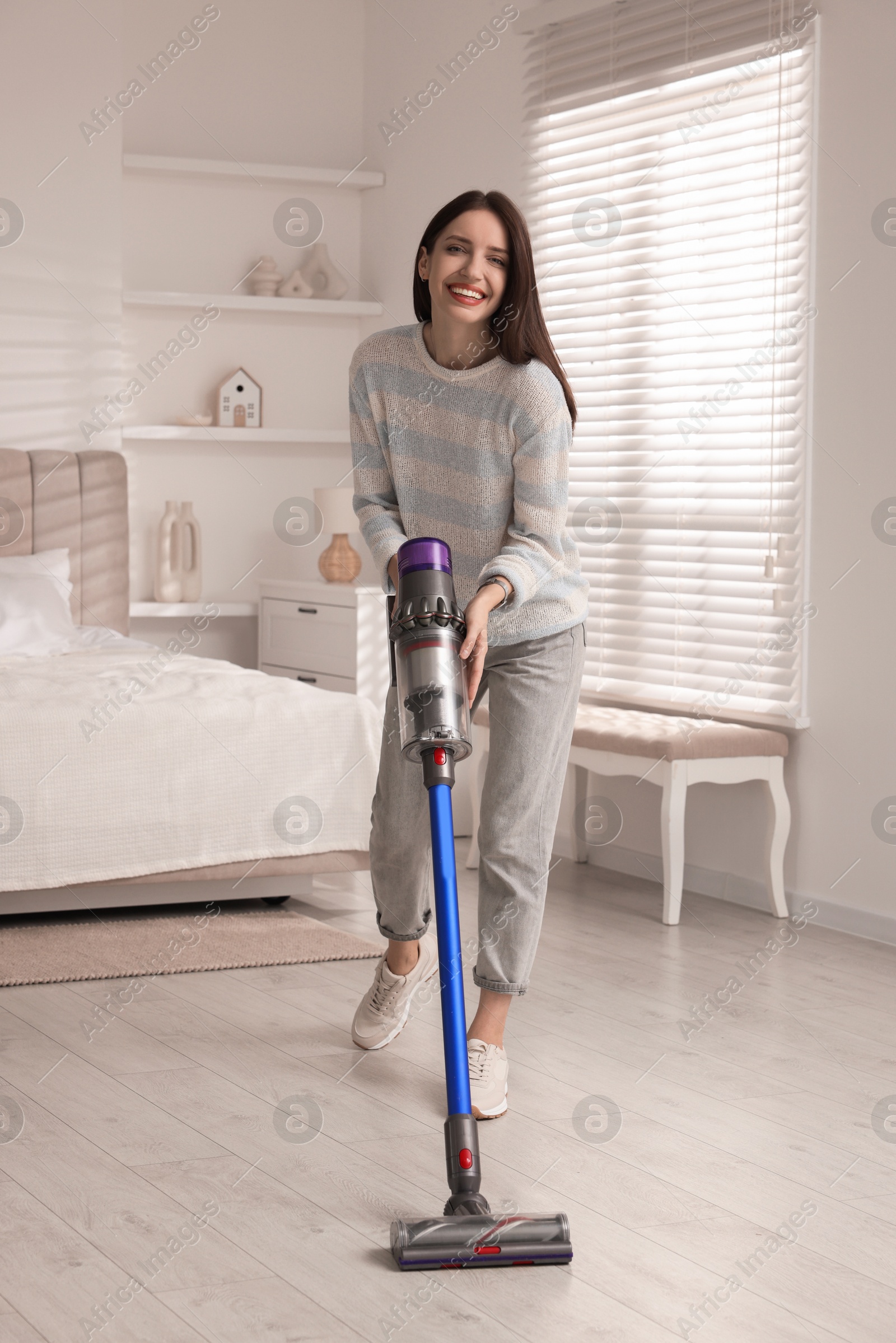 Photo of Smiling young woman cleaning floor with cordless vacuum cleaner in bedroom