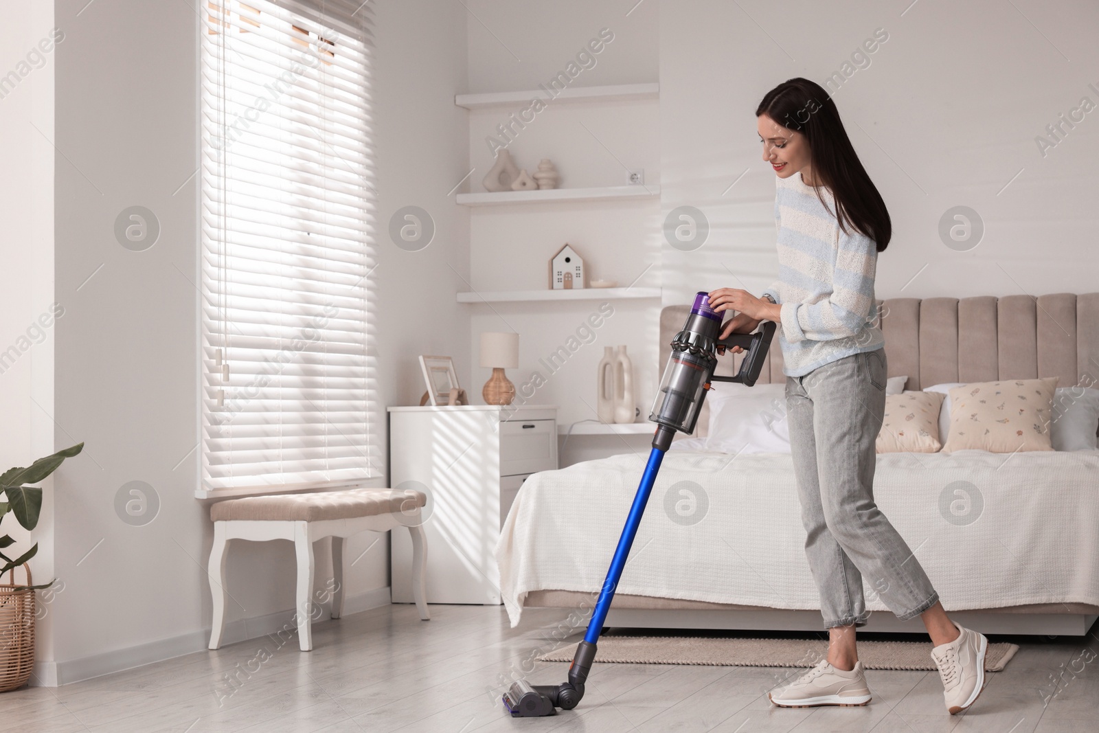 Photo of Young woman cleaning floor with cordless vacuum cleaner in bedroom