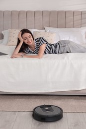 Photo of Smiling young woman resting on bed while robotic vacuum cleaner vacuuming floor at home