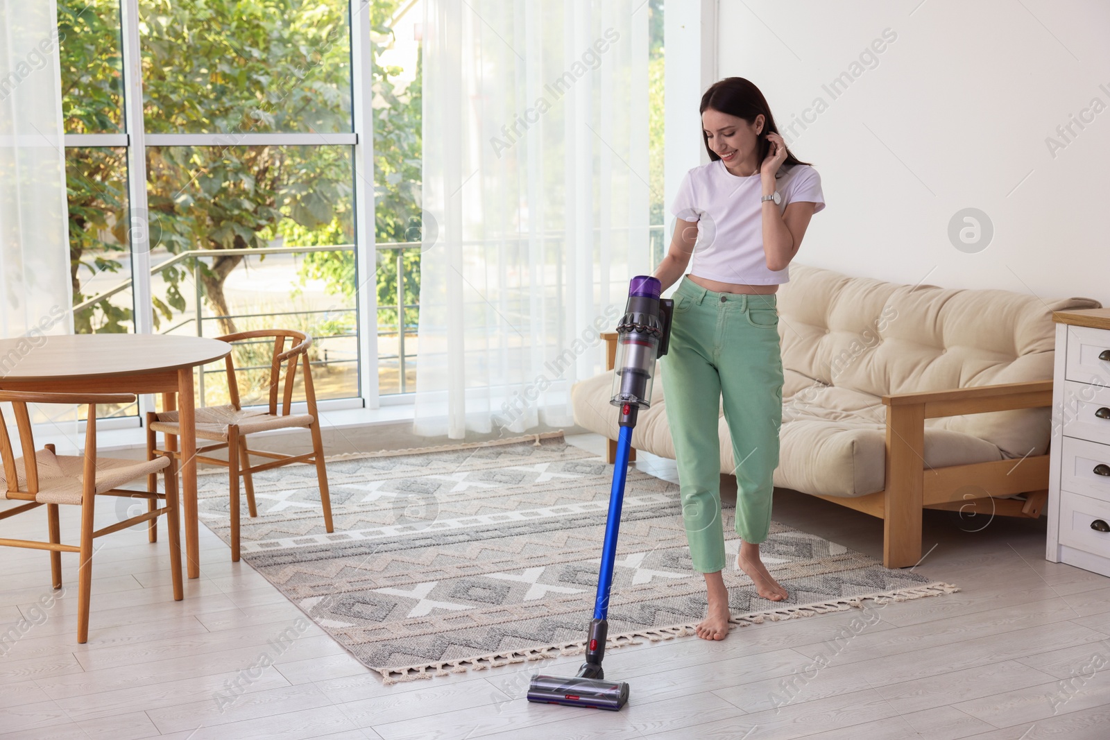 Photo of Smiling young woman cleaning floor with cordless vacuum cleaner at home