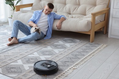 Photo of Young man resting with his Neva Masquerade cat while robotic vacuum cleaner vacuuming rug at home