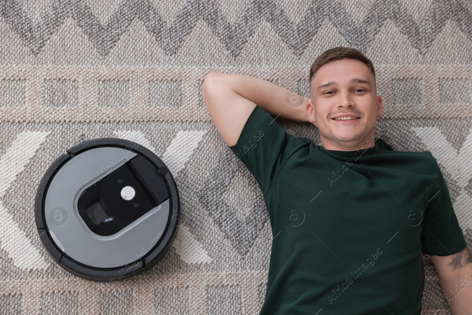 Photo of Smiling young man and robotic vacuum cleaner on rug, top view