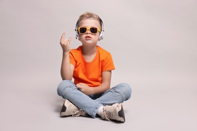 Little boy with sunglasses listening to music and showing rock gesture on light grey background