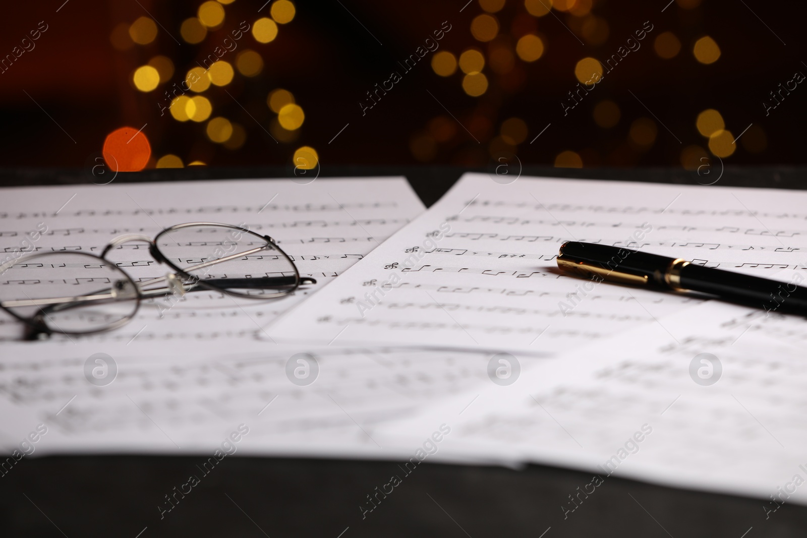 Photo of Sheets of musical notes and pen on table against blurred lights, closeup. Bokeh effect