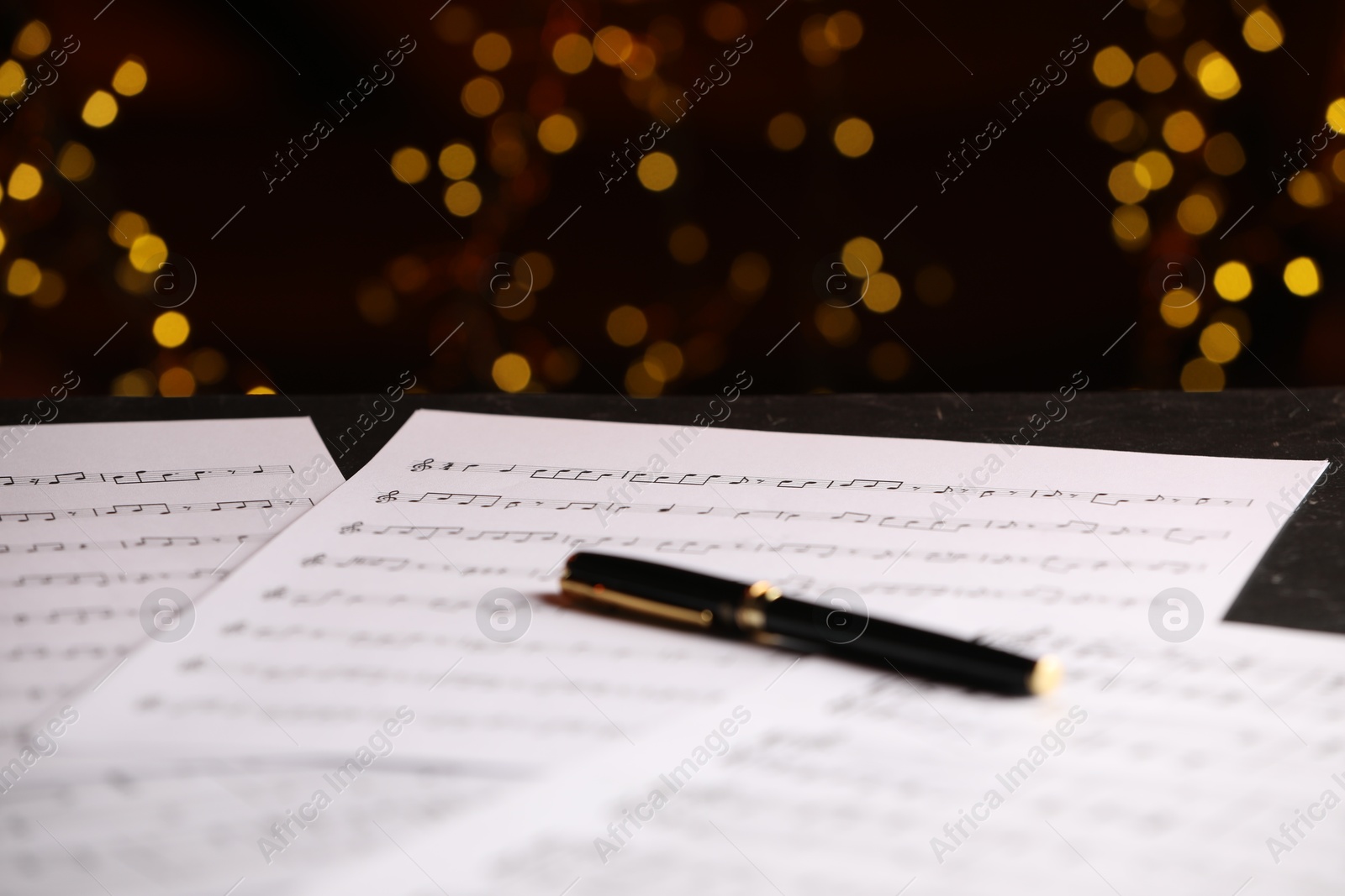 Photo of Sheets of musical notes and pen on table against blurred lights, closeup. Bokeh effect