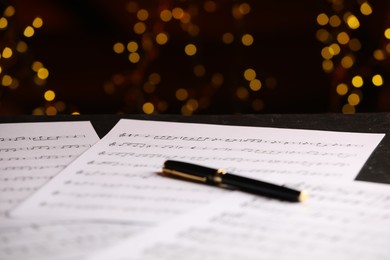 Photo of Sheets of musical notes and pen on table against blurred lights, closeup. Bokeh effect