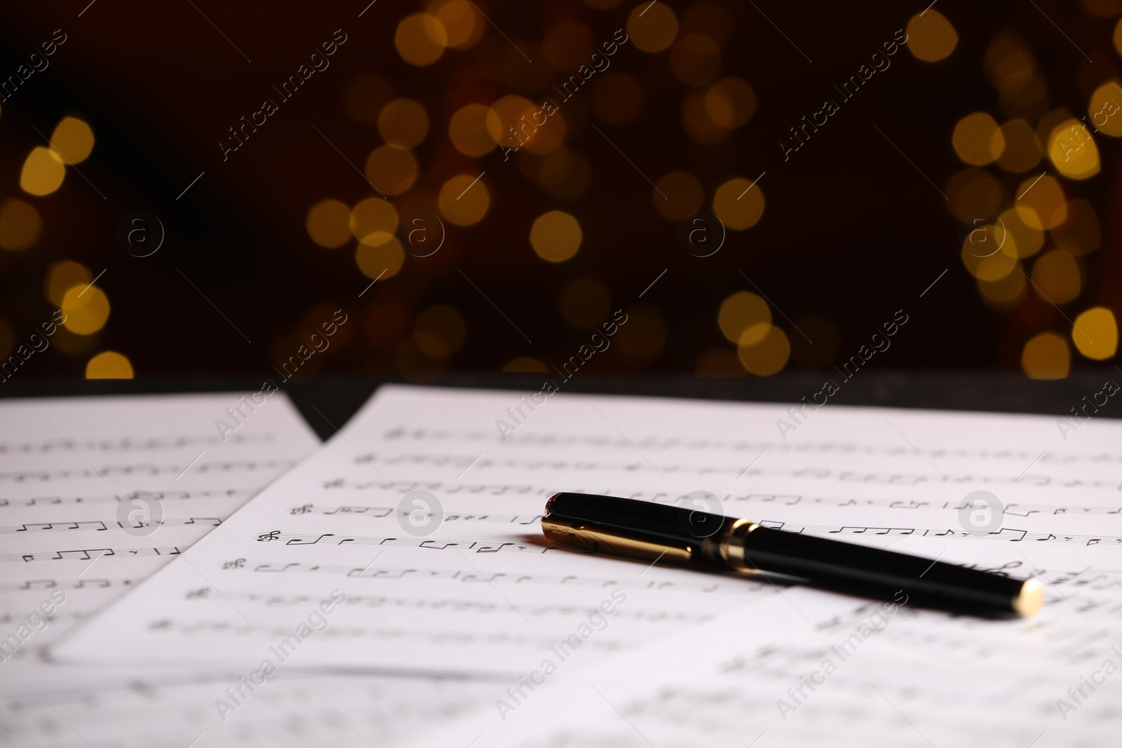 Photo of Sheets of musical notes and pen on table against blurred lights, closeup. Bokeh effect