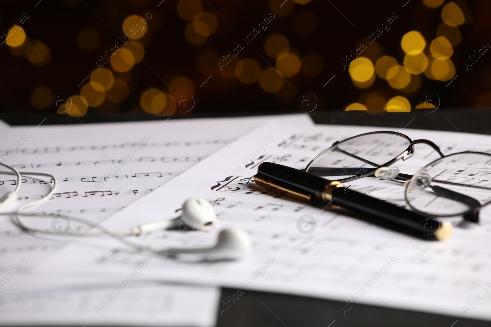 Photo of Sheets of musical notes, glasses, earphones and pen on table against blurred lights, closeup. Bokeh effect