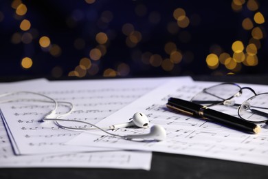 Photo of Sheets of musical notes, glasses, earphones and pen on table against blurred lights, closeup. Bokeh effect