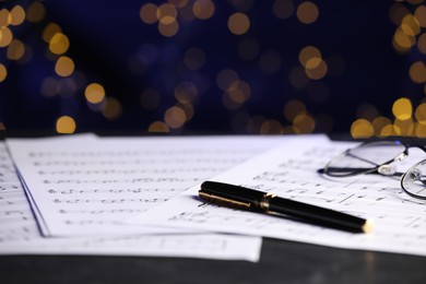 Photo of Sheets of musical notes, glasses and pen on table against blurred lights, closeup. Bokeh effect