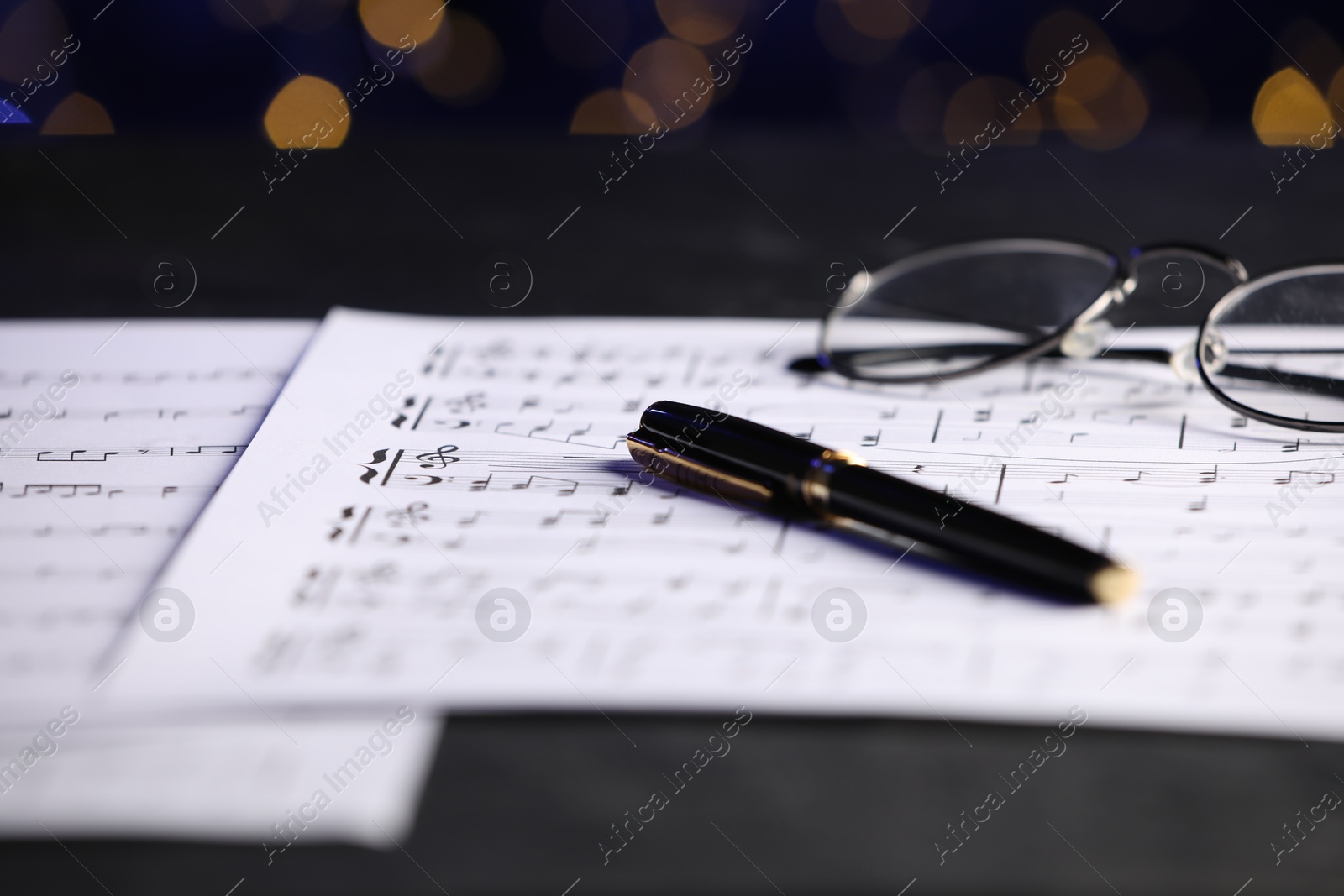 Photo of Sheets of musical notes, glasses and pen on table against blurred lights, closeup. Bokeh effect