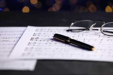 Photo of Sheets of musical notes, glasses and pen on table against blurred lights, closeup. Bokeh effect