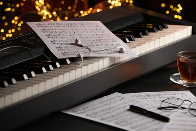 Photo of Piano with musical notes, earphones, glasses and cup of tea on table against blurred lights, closeup. Bokeh effect