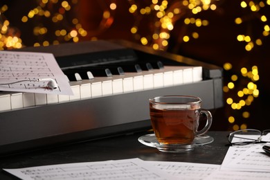 Photo of Piano with musical notes, earphones and cup of tea on table against blurred lights, closeup. Bokeh effect