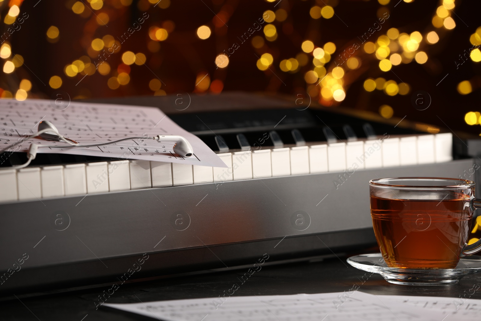 Photo of Piano with musical notes, earphones and cup of tea on table against blurred lights, closeup. Bokeh effect