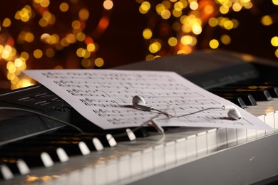 Photo of Sheet with musical notes and earphones on piano against blurred lights, closeup. Bokeh effect