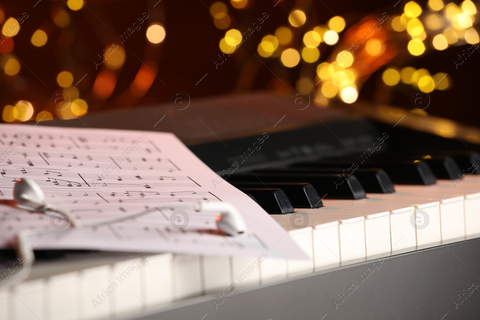 Photo of Sheet with musical notes and earphones on piano against blurred lights, closeup. Bokeh effect