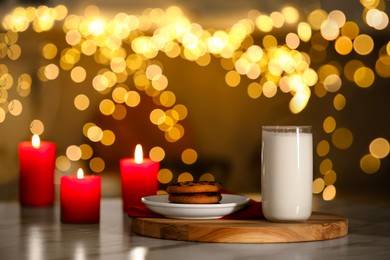 Photo of Glass of milk, tasty chocolate chip cookies and burning candles on table against blurred Christmas lights. Bokeh effect