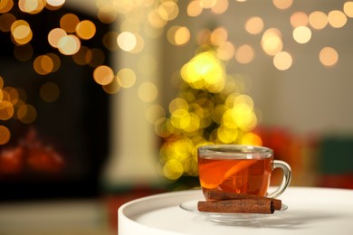 Photo of Cup of tea with lemon and cinnamon sticks on table against blurred Christmas lights, bokeh effect. Space for text