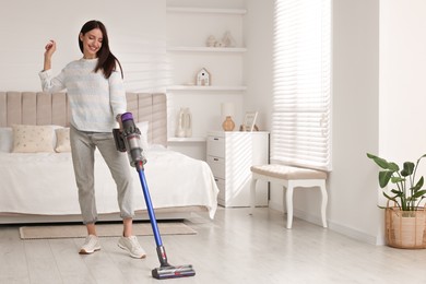 Photo of Smiling young woman cleaning floor with cordless vacuum cleaner in bedroom