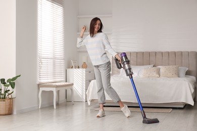 Photo of Smiling young woman cleaning floor with cordless vacuum cleaner in bedroom