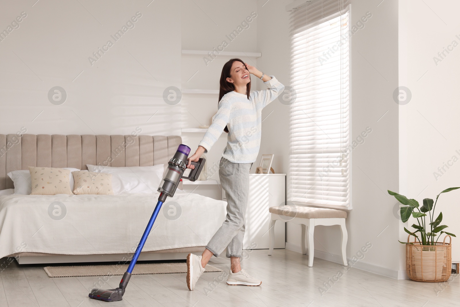 Photo of Smiling young woman cleaning floor with cordless vacuum cleaner in bedroom