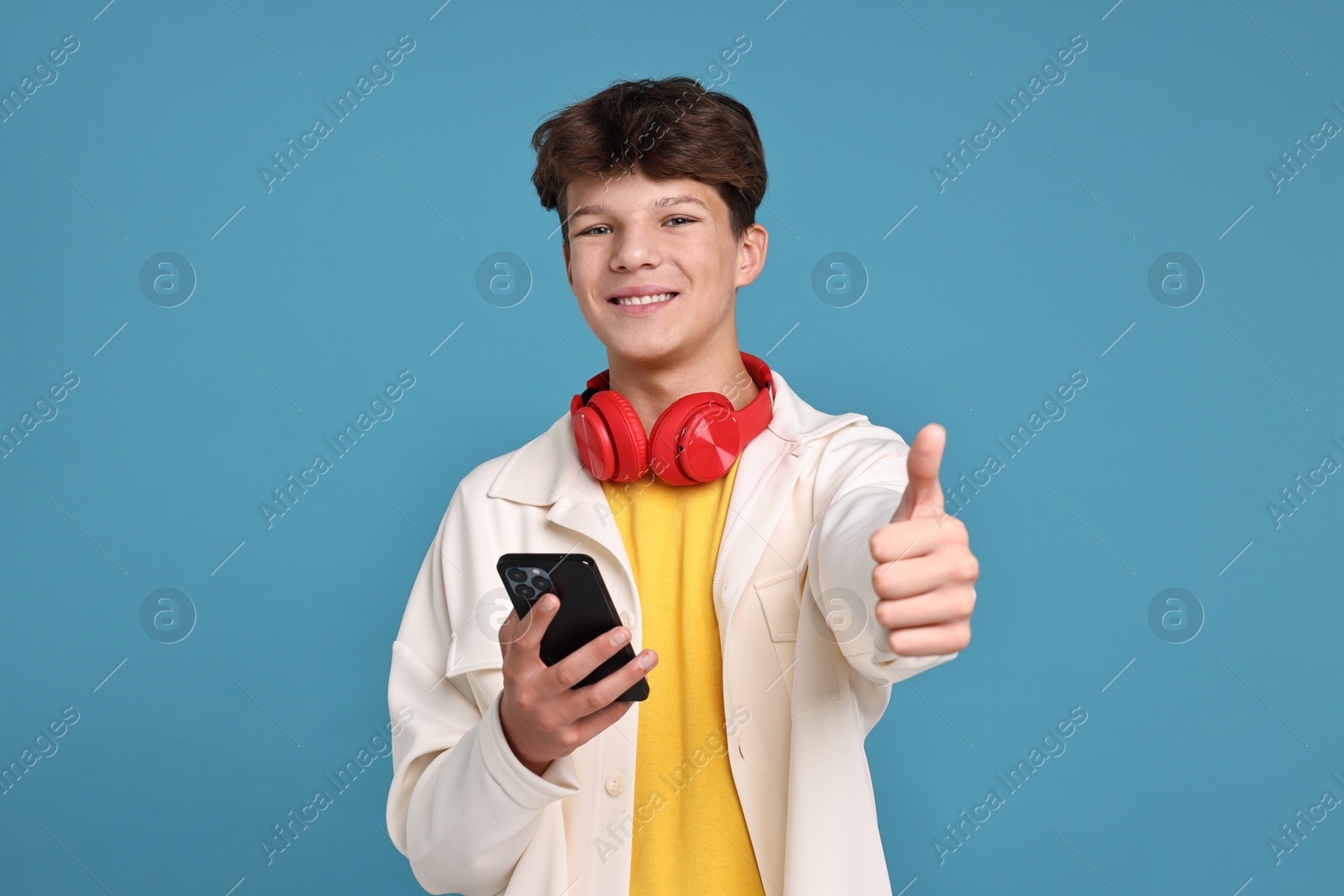 Photo of Teenage boy with headphones and smartphone showing thumbs up on light blue background