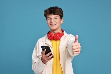 Photo of Teenage boy with headphones and smartphone showing thumbs up on light blue background