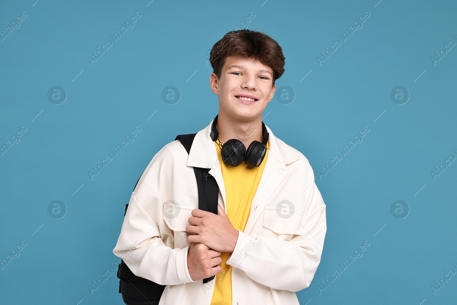 Photo of Happy teenage boy with headphones and backpack on light blue background