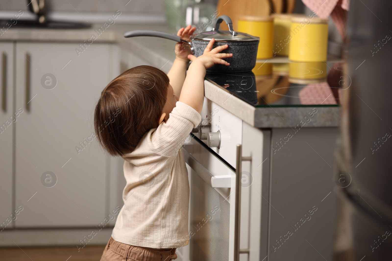 Photo of Little boy playing with pot on stove in kitchen. Dangerous situation