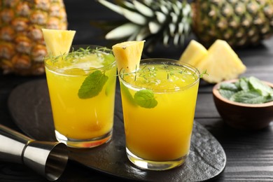 Photo of Tasty pineapple cocktail with mint in glasses and jigger on black wooden table, closeup