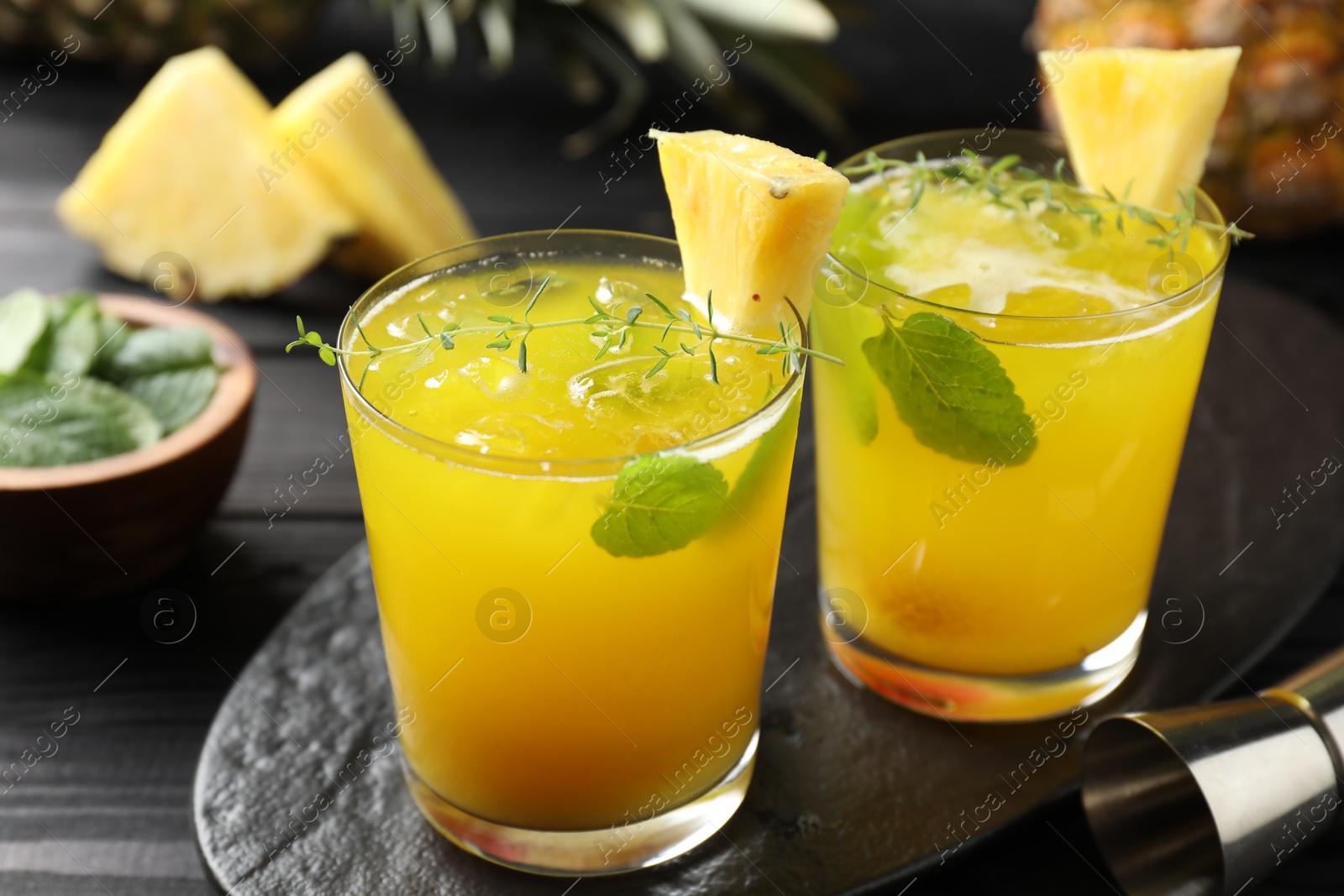 Photo of Tasty pineapple cocktail with mint in glasses and jigger on black wooden table, closeup