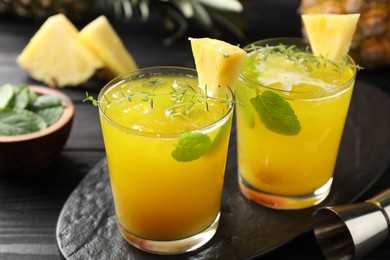 Photo of Tasty pineapple cocktail with mint in glasses and jigger on black wooden table, closeup