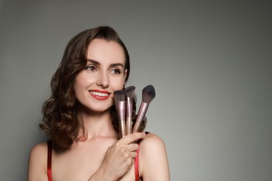 Photo of Smiling woman with different makeup brushes on grey background. Space for text