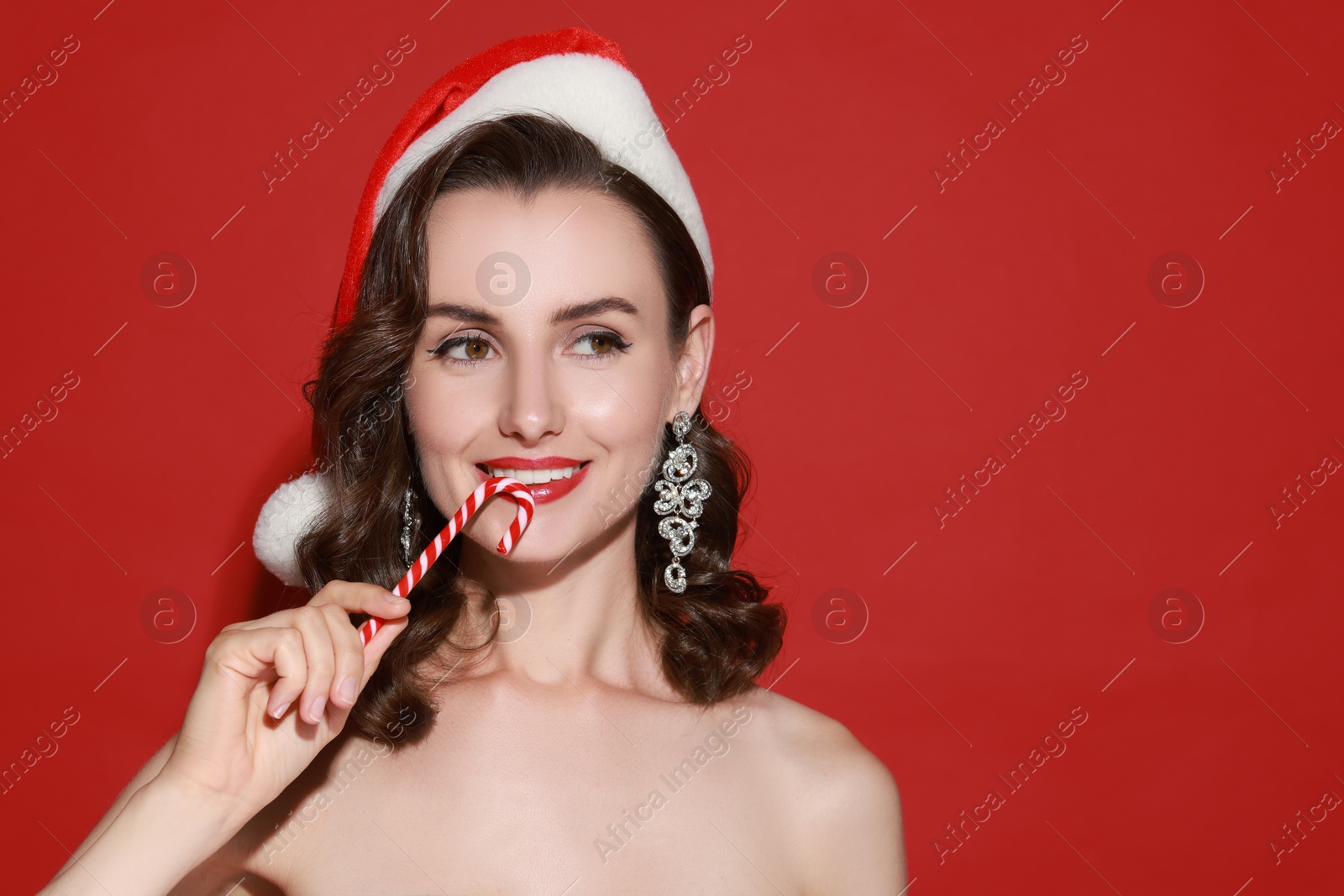 Photo of Smiling woman with perfect makeup in Santa hat holding Christmas candy cane on red background. Space for text