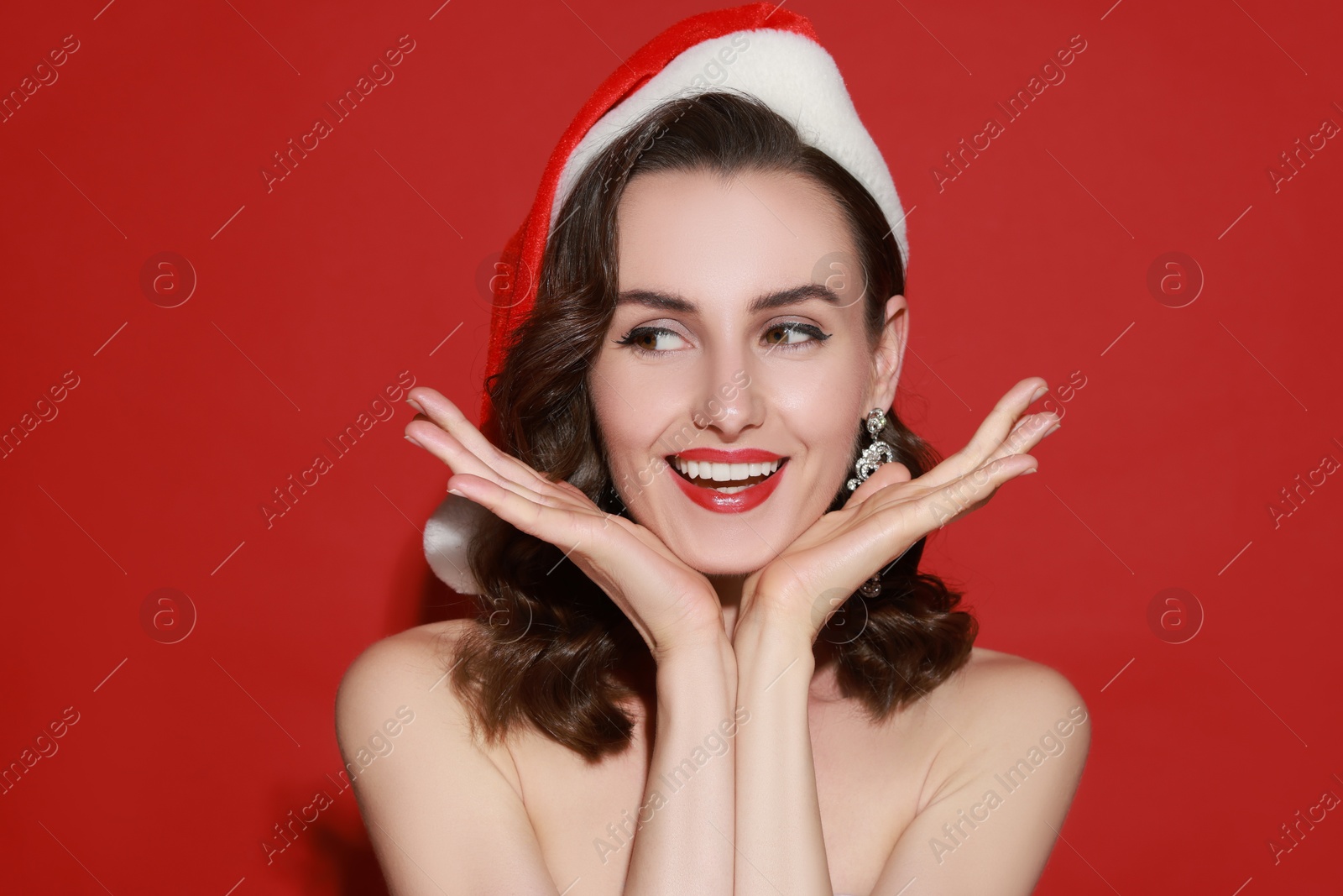 Photo of Smiling woman with perfect makeup in Santa hat on red background. Christmas celebration