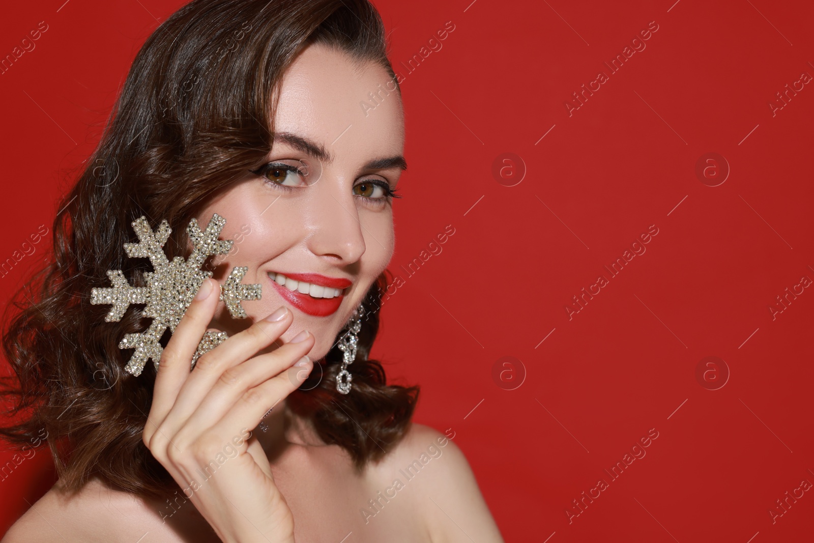 Photo of Smiling woman with perfect makeup holding decorative snowflake on red background, space for text. Christmas celebration