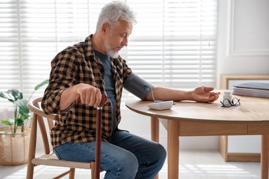 Photo of Senior man with walking cane measuring blood pressure at table indoors