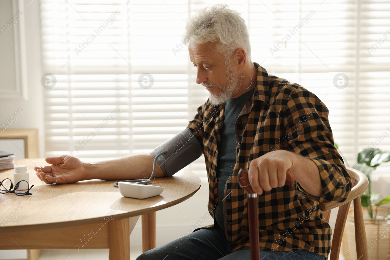 Photo of Senior man with walking cane measuring blood pressure at table indoors