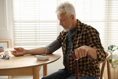 Photo of Senior man with walking cane measuring blood pressure at table indoors