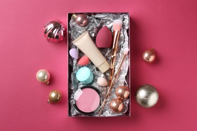 Photo of Christmas present. Decorative cosmetics in gift box and festive balls on dark pink background, top view