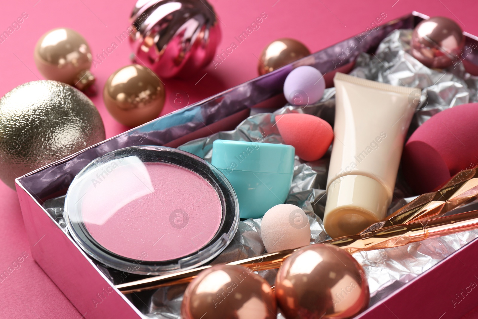 Photo of Christmas present. Decorative cosmetics in gift box and festive balls on dark pink background, closeup