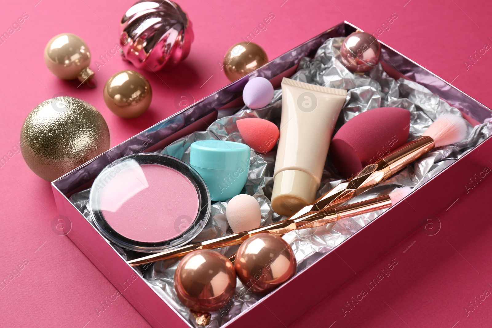 Photo of Christmas present. Decorative cosmetics in gift box and festive balls on dark pink background, closeup