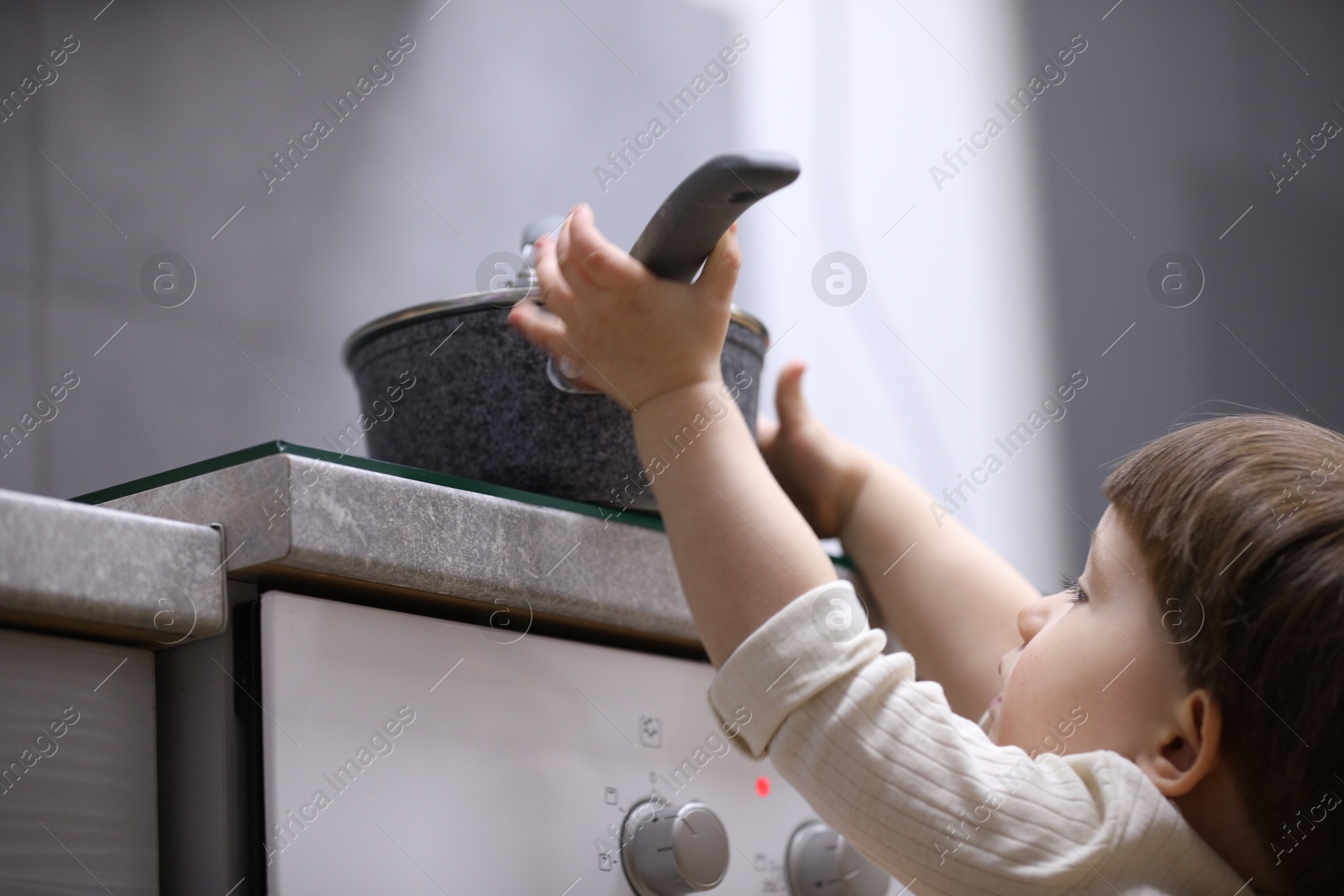 Photo of Little boy playing with pot on stove in kitchen, low angle view. Dangerous situation