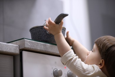 Photo of Little boy playing with pot on stove in kitchen, low angle view. Dangerous situation