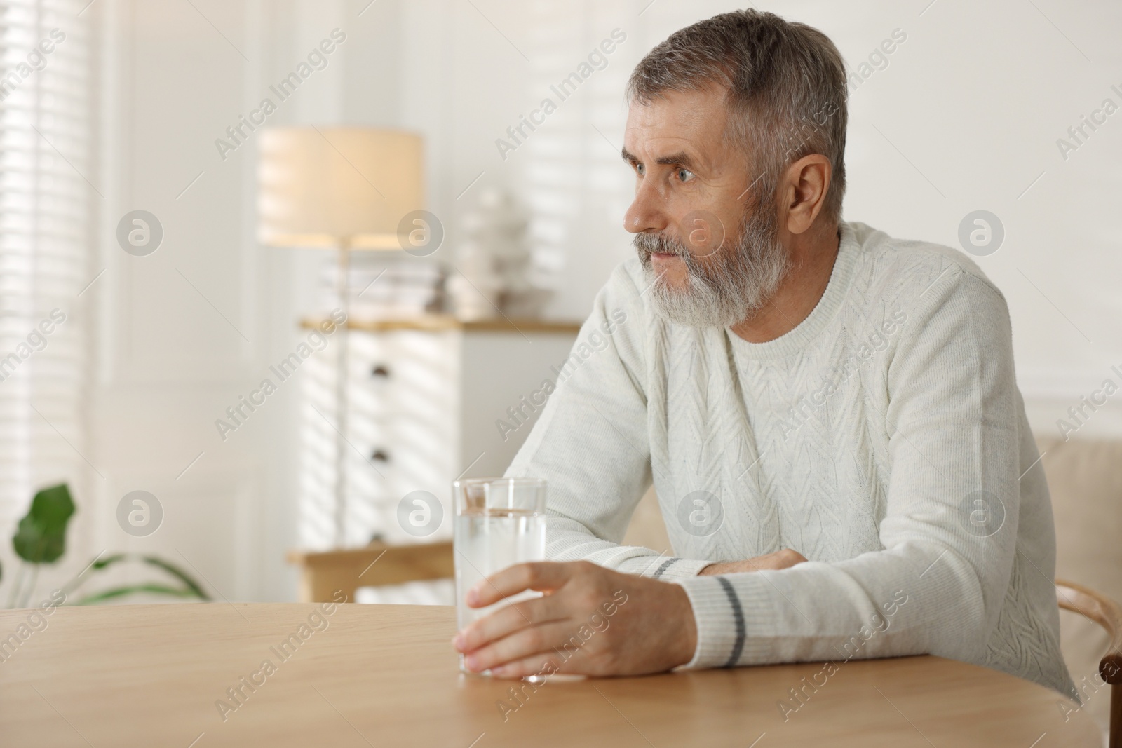 Photo of Senior man with glass of water at table indoors, space for text