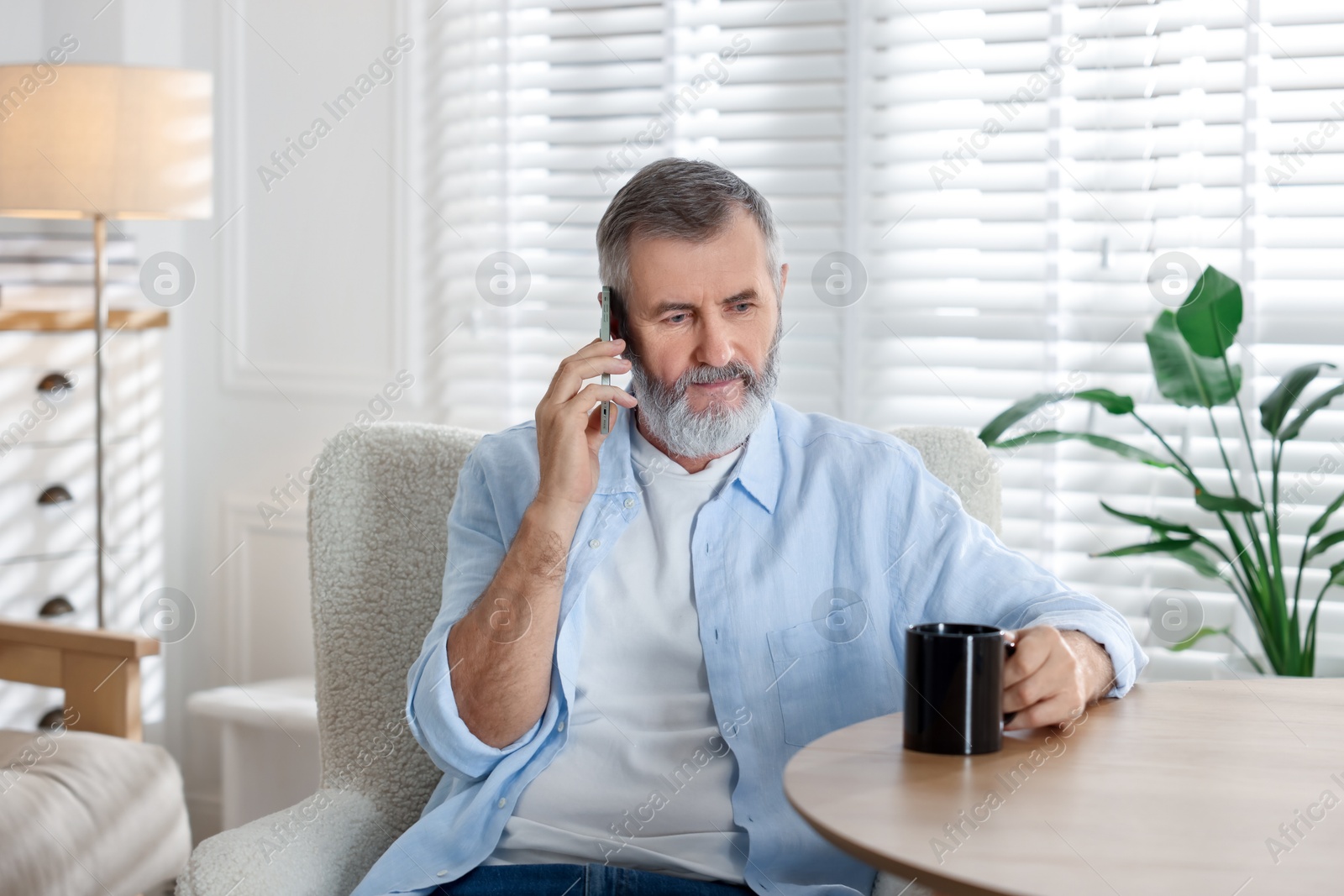 Photo of Senior man with cup of drink talking on smartphone at table indoors