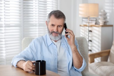 Photo of Senior man with cup of drink talking on smartphone at table indoors