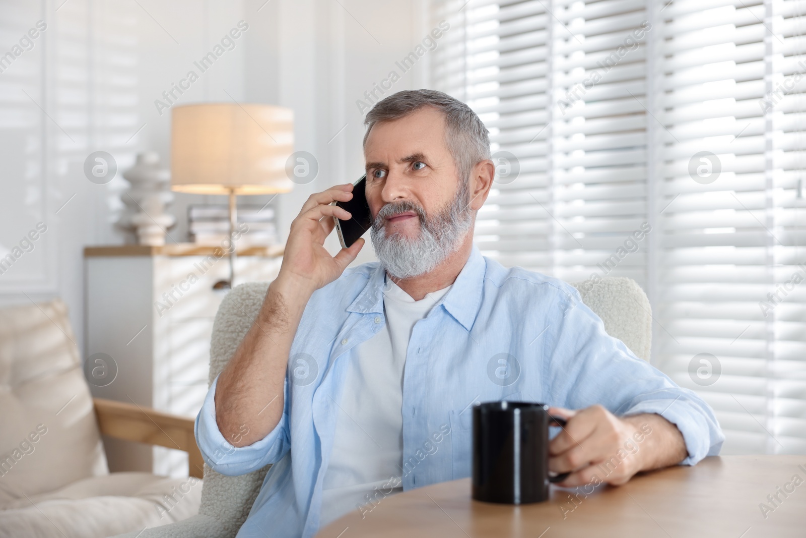 Photo of Senior man with cup of drink talking on smartphone at table indoors